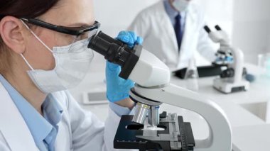 Maskeli profesyonel kadın bilim adamı mikroskop ile numune inceleyerek steril laboratuvar ortamında meslektaşlarıyla birlikte araştırma yapıyor. Tıp, sağlık ve bilim konsepti.