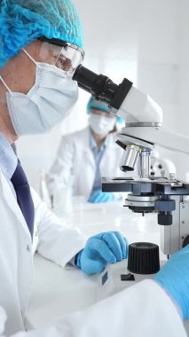Koruyucu maske, şapka ve eldiven takan kıdemli erkek bilim adamı modern bir laboratuvarda mikroskop altında örnekleri inceliyor. Tıp, sağlık ve bilim konsepti.