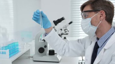 Koruyucu giysi giyen bilim araştırmacısı modern laboratuvar çalışma alanında mavi sıvı numunesini test tüpünden inceliyor. Tıp, sağlık ve bilim konsepti