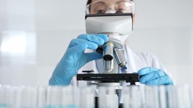 Koruyucu ekipman takan kadın bilim adamı mikroskobu kontrol ediyor, modern laboratuvar araştırma ortamında mavi test tüplerini analiz ediyor. Tıp, sağlık ve bilim konsepti.