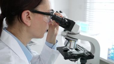 Laboratuvarda mikroskop kullanarak laboratuvar önlüğü ve güvenlik gözlüğü takan kadın bilim adamı. Arka planda deney tüpleri ile bilimsel araştırma yapıyor. Tıp, sağlık ve bilim konsepti