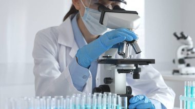 Beyaz koruyucu giysili kadın bir bilim adamı mikroskop merceğini ayarlar ve steril bir laboratuvar ortamında tıbbi araştırma örneklerini analiz eder..