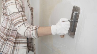 Eldiven giyen kimliği belirsiz kadın inşaat işçisi duvara sıva uygulamak için macun bıçağı kullanıyor. Ev yenileme projesi sırasında yüzeyi düzleştiriyor.