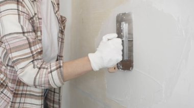 Eldiven giyen kimliği belirsiz kadın inşaat işçisi duvara sıva uygulamak için macun bıçağı kullanıyor. Ev yenileme projesi sırasında yüzeyi düzleştiriyor.