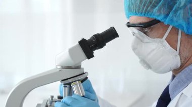 Koruyucu ekipman takan erkek bilim adamı mikroskop kaydırağını inceliyor, steril laboratuvar ortamında araştırma örneklerini analiz ediyor. Sağlık ve bilim konsepti.
