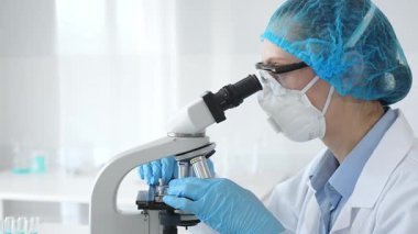 Kadın araştırmacı mikroskop ile numune inceliyor, steril laboratuvar içinde çalışma yerini çevreleyen cam şişelerle tıbbi deney yapıyor. Tıp ve Bilim.