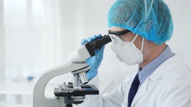 Üst düzey erkek bilim adamı koruyucu maske, saç filesi, gözlük ve eldiven takıyor mikroskop kullanarak tıbbi bir laboratuarda bilimsel araştırma yapıyor. Laboratuvar ve bilim konsepti