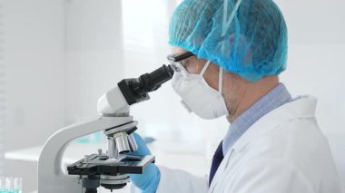 Üst düzey erkek bilim adamı koruyucu maske, saç filesi, gözlük ve eldiven takıyor mikroskop kullanarak tıbbi bir laboratuarda bilimsel araştırma yapıyor. Laboratuvar ve bilim konsepti