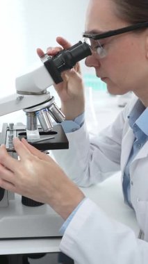 Laboratuvar önlüğü ve koruyucu gözlüklü kadın bilim adamı modern bir laboratuvarda mikroskop altında test örnekleri inceliyor. Tıp, sağlık ve bilim konsepti
