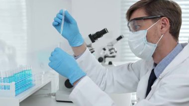 Laboratuvar önlüklü, koruyucu gözlüklü ve mavi eldivenli maskeli erkek bilim adamı modern laboratuardaki test tüplerine mikroskop ve test tüpleri rafı koyuyor. Tıp, sağlık ve bilim konsepti.