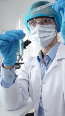 Maske takan kadın bilim adamı, mavi başlık ve eldivenler laboratuardaki pipeti kullanarak test tüpüne sıvı ekliyor. Tıp ve bilim konsepti.