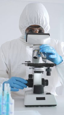 Koruyucu giysi giyen tıp bilimcisi örnekleri incelerken mikroskop kullanıyor steril laboratuvar ortamında araştırma yapıyor. Mavi sıvıyla test tüplerinin yakın plan görüntüsü.