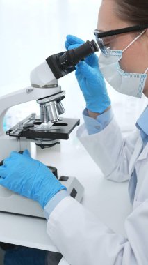 Mavi koruyucu giysili kadın bilim adamı laboratuvar araştırma ortamında test örneklerini mikroskop altında analiz ediyor. Tıp, sağlık ve bilim konsepti.