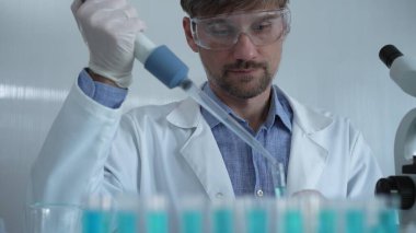 Laboratuvar önlüğü ve güvenlik gözlüğü takan bilim adamı deney tüpüne sıvı atmak için mikropipet kullanıyor. Portre görüntüsü. Tıp ve Bilim.