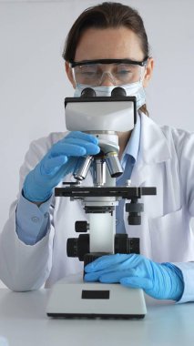 Mikroskop kaydırağını inceleyen kadın bilim adamı, laboratuarda koruyucu giysiler giyiyor, mavi renkli test tüpleriyle farmasötik buluşları araştırıyor..