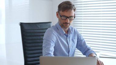 Mavi parlak gömlek ve siyah gözlük giyen profesyonel muhasebeci, parlak ve modern bir ofiste çalışırken profesyonelliğini gösteriyor. İş adamları konsepti.