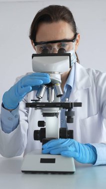 Mikroskop kaydırağını inceleyen kadın bilim adamı, laboratuarda koruyucu giysiler giyiyor, mavi renkli test tüpleriyle farmasötik buluşları araştırıyor..