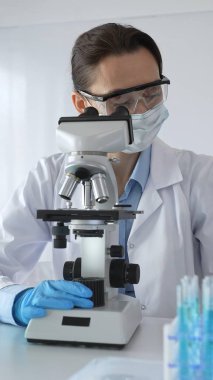 Koruyucu gözlük takan ve maske takan kadın bilim adamı mikroskop kullanarak laboratuvarda numuneleri analiz ediyor mavi sıvı test tüpleriyle çevrili yeni ilaç veya aşıları araştırıyor.
