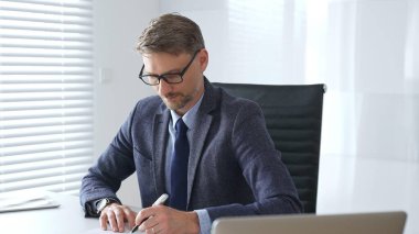 Kıdemli işadamı takım elbise, kuyruk ve siyah gözlük takarak masasındaki parlak, modern bir ofiste profesyonelliği gösteren bir belgeye konsantre oluyor. İş adamları konsepti.