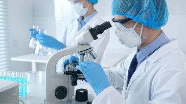 Koruyucu maske takan ve gözlük takan bilim adamı mikroskop kullanarak laboratuvarda araştırma yapıyor ve arka planda elinde mataralar olan bir bilim adamı asistanı var. Sağlık ve bilim konsepti.