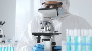 Koruyucu kıyafetli ve eldivenli bir erkek doktor örnekleri incelerken mikroskop kullanır ve laboratuvarda araştırma yapar. Mavi sıvıyla test tüplerinin yakın plan görüntüsü.