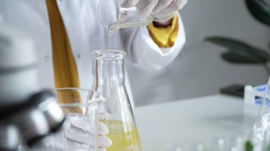 Laboratuvar önlüğü ve beyaz eldiven giyen bir bilim adamı laboratuvar ortamında sarı yağlı sıvıyı bir bardaktan diğerine döküyor. Bilim ve tıp.