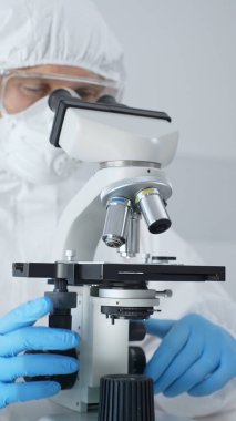 Tehlikeli madde giysisi, maske ve mavi eldiven giyen erkek bir virüs bilimci laboratuvarda virüsleri araştırırken mikroskop altında özenle çalışıyor. Tıp ve bilim konsepti.