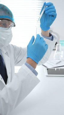 Laboratuvar şapkası, yüz maskesi, Google ve mavi eldiven takan erkek tıp bilimci modern bir laboratuvar ortamında bir deney tüpünün içine dikkatlice sıvı bırakıyor. Tıp, sağlık hizmeti kavramı.