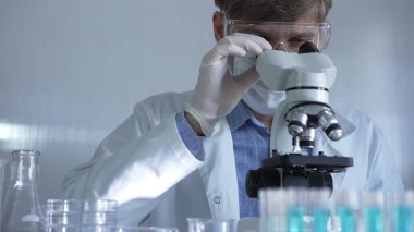 Laboratuvar önlüğü, maskesi, beyaz eldivenleri ve güvenlik gözlüğü takan erkek bilim adamı laboratuarda test tüpleriyle çevrili bir dürbün kullanıyor..