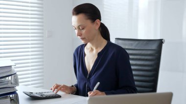 Masada hesap makinesiyle oturan ve not alan, modern ofis ortamında finansal verileri ve evrak işlerini yöneten odaklı bir iş kadını. İş dünyasında denetim ve vergi teması