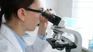 Laboratuvarda mikroskop kullanarak laboratuvar önlüğü ve güvenlik gözlüğü takan kadın bilim adamı. Arka planda deney tüpleri ile bilimsel araştırma yapıyor. Tıp, sağlık ve bilim konsepti