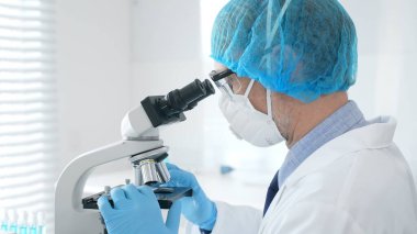 Tehlikeli madde giysisi, maskesi ve eldiveni olan bir tıp virologu bir laboratuvar araştırma virüsünün mikroskobunda özenle çalışıyor. Sağlık ve Bilim Konsepti.