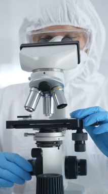 Tehlikeli madde giysisi, maskesi ve mavi eldivenli bir erkek virüs uzmanı laboratuvar araştırmalarında mikroskop başında özenle çalışıyor. Tıp, bilim konsepti.