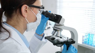 Kadın araştırmacı mikroskop ile örnek analiz ediyor, koruyucu teçhizat takılı, steril araştırma ortamında gelişmiş laboratuvar ekipmanlarının yanında çalışan bir meslektaşı. Tıp, sağlık ve bilim konsepti
