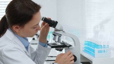 Modern bir laboratuvarda mikroskopla çalışan kadın bilim adamı mavi sıvı içeren test tüplerindeki örnekleri analiz ediyor ve bilimsel araştırmalar yürütüyor. Tıp, sağlık ve bilim konsepti