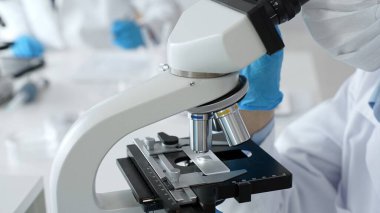 Mavi eldiven ve maske takan bilim adamları, laboratuvar ortamında modern bir mikroskop kullanarak araştırma ve analiz yapıyorlar. Tıp, sağlık ve bilim konsepti