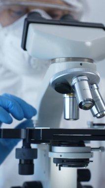 Tehlikeli madde giysisi, maskesi ve eldivenleri olan bir tıp virologu laboratuvar araştırmalarında mikroskop başında özenle çalışıyor. Tıp ve bilim konsepti.