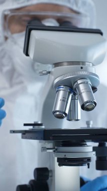 Tehlikeli madde giysisi, maskesi ve eldiveni olan bir tıp virologu bir laboratuvar araştırma virüsünün mikroskobunda özenle çalışıyor. Sağlık ve Bilim Konsepti.