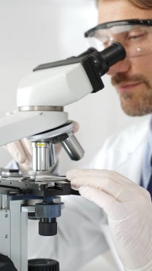 Erkek araştırmacının mikroskopla incelediği örnek, steril laboratuvar ortamında güvenlik gözlüğü takıyor, bilimsel cam eşyalar ile çevrili. Tıp, sağlık ve bilim konsepti.