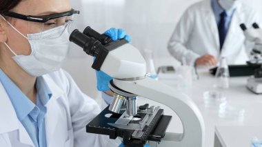 Kadın araştırmacı mikroskop ile örnek analiz ediyor, koruyucu teçhizat takılı, steril araştırma ortamında gelişmiş laboratuvar ekipmanlarının yanında çalışan bir meslektaşı. Tıp, sağlık ve bilim konsepti