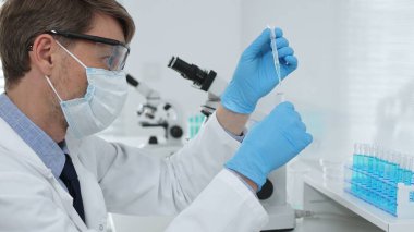 Laboratuvar önlüklü, koruyucu gözlüklü ve mavi eldivenli maskeli erkek bilim adamı modern laboratuardaki test tüplerine mikroskop ve test tüpleri rafı koyuyor. Tıp, sağlık ve bilim konsepti.