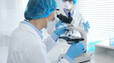 Mavi koruyucu donanımlı kadın bilim adamı mikroskop altında numuneleri incelerken laboratuvar asistanı da arka planda cam parçalarıyla ilgileniyor. Tıp ve bilim konsepti.
