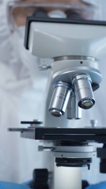 Tehlikeli madde giysisi, maskesi ve eldivenleri olan bir tıp virologu laboratuvar araştırmalarında mikroskop başında özenle çalışıyor. Tıp ve bilim konsepti.