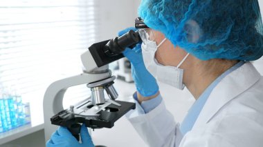 Maske takan ve mavi güvenlik donanımı takan kadın bir bilim adamı araştırma için mikroskop kullanırken erkek bir meslektaşı arka planda çalışıyor. Tıp, sağlık ve bilim konsepti.