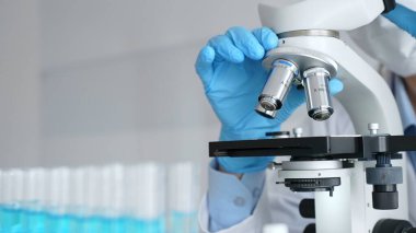 Mikroskobu ayarlayan eldivenli bilim adamlarına yakın çekim yapmak, laboratuvarda araştırma yapmak ve arka planda mavi sıvı içeren test tüpleri kullanmak. Tıp, sağlık ve bilim konsepti.
