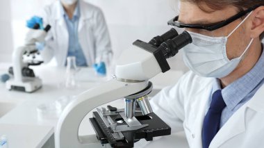 Mikroskop kullanarak maske takan ve eldiven takan bilim adamlarına yakın çekim, modern laboratuvarda araştırmalar yürütmek ve arka planda çalışmak. Tıp, sağlık ve bilim konsepti