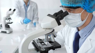 Koruyucu maske, şapka ve eldiven takan kıdemli erkek bilim adamı modern bir laboratuvarda mikroskop altında örnekleri inceliyor. Tıp, sağlık ve bilim konsepti.