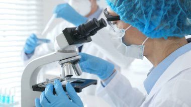Mikroskop altında numuneleri incelerken mavi koruyucu giysili kadın bilim adamlarının yakın plan çekimlerinde laboratuvar asistanları arka planda cam parçaları incelerler. Tıp konsepti.