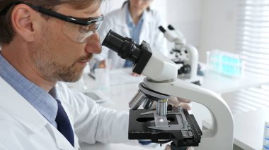 Koruyucu gözlük takan bilim adamı mikroskop altında araştırma ve analiz yapıyor. Arka planda test tüpleri ile modern bir laboratuvarda. Tıp, sağlık ve bilim konsepti