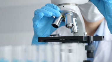 Mikroskobu ayarlayan eldivenli bilim adamlarına yakın çekim yapmak, laboratuvarda araştırma yapmak ve arka planda mavi sıvı içeren test tüpleri kullanmak. Tıp, sağlık ve bilim konsepti.